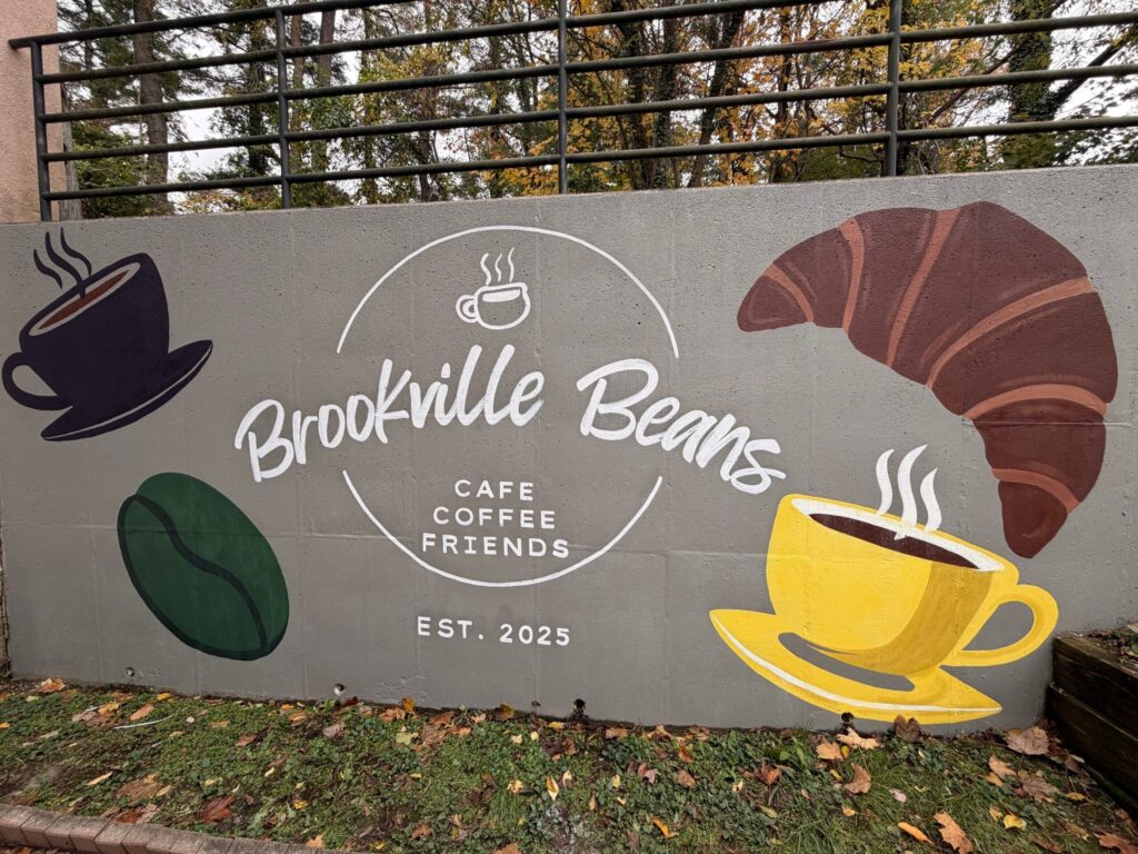 A painted wall reads "Brookville Beans Cafe Coffee Friends Est. 2025" with paintings of coffee cups, a croissant and a coffee bean.