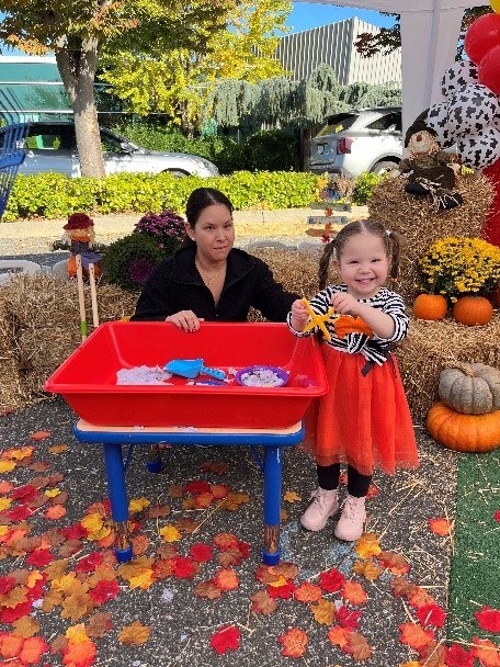 A Barbara C. Wilson Preschool student and a teacher smile while playing with a sensory table filled with sand at the Fall Festival.