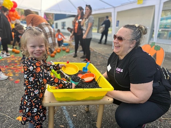 A Barbara C. Wilson Preschool student and a teacher smile while playing with a sensory table filled with beans at the Fall Festival.