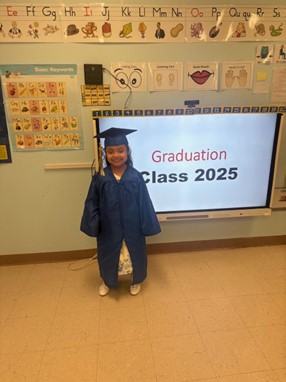 A BCCS graduate stands in front of a screen that reads "Graduation Class 2025"