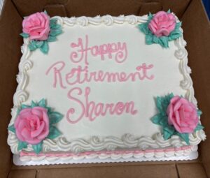 A white cake with "Happy Retirement Sharon" written in pink frosting
