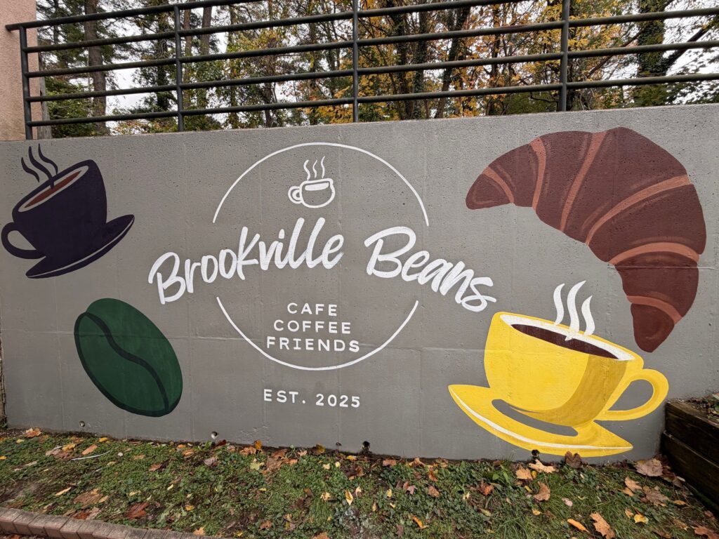 A painted wall reads "Brookville Beans Cafe Coffee Friends Est. 2025" with paintings of coffee cups, a croissant and a coffee bean.